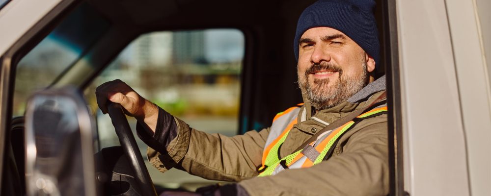 Smiling truck driver looking through side window while driving his truck and looking at camera.